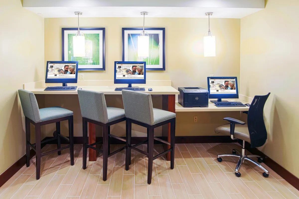 A productive business center setup with several computer stations, tasteful artwork, and ample lighting. Guests are seen catching up on work, printing documents, and interacting in a collaborative, professional environment.