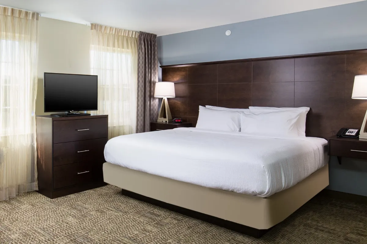 A warmly lit bedroom featuring a plush king-sized bed with crisp linens, nightstands with modern lamps, a wall-mounted television, and a roomy dresser. Natural light streams in from a window, creating an inviting, restful atmosphere ideal for relaxation and sleep.
