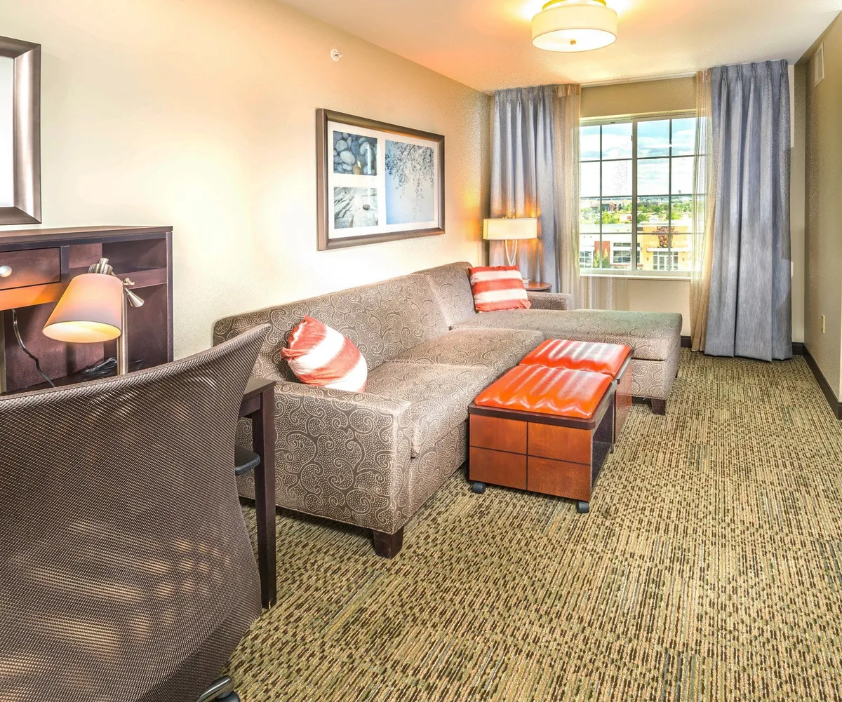 A sunlit suite interior featuring a plush sofa, modern coffee table, work desk, and large window inviting in cheerful morning light. The inviting space is perfect for relaxing, catching up on work, or enjoying a quiet moment with a book or coffee.