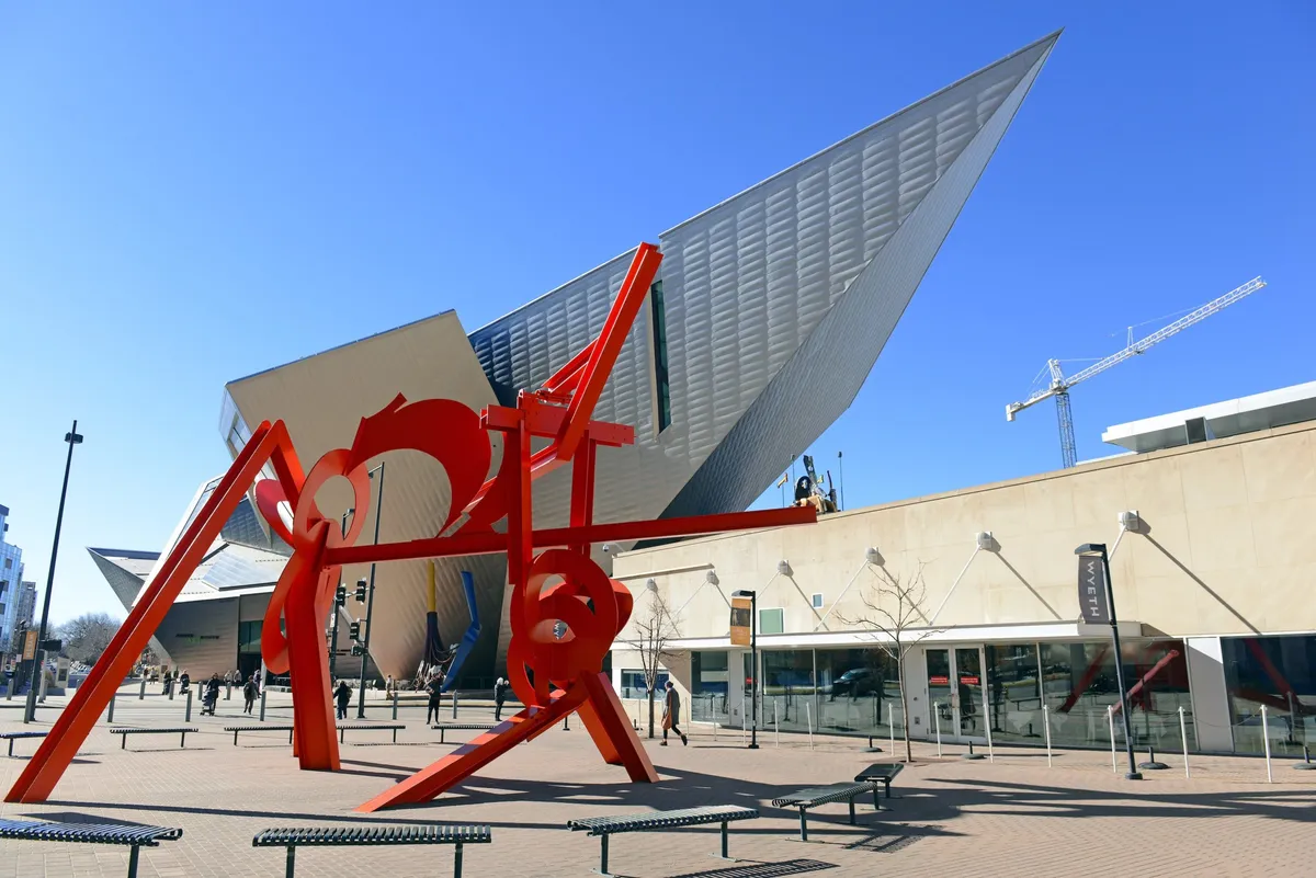 A striking red sculpture stands in front of a modern building, with benches and people gathering in the surrounding plaza. The scene conveys Denver's artistic energy and inviting public spaces, ideal for taking a quick break or meeting colleagues outside.