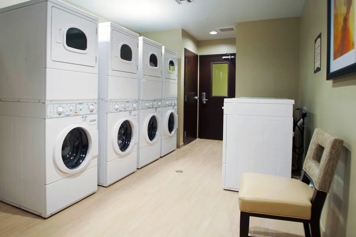 The image features a clean and practical laundry area with stacked washer and dryer units, a single washing machine, and a small seating area for guests waiting to finish their laundry. The efficient arrangement and tidy appearance reflect both convenience and attention to guest needs.