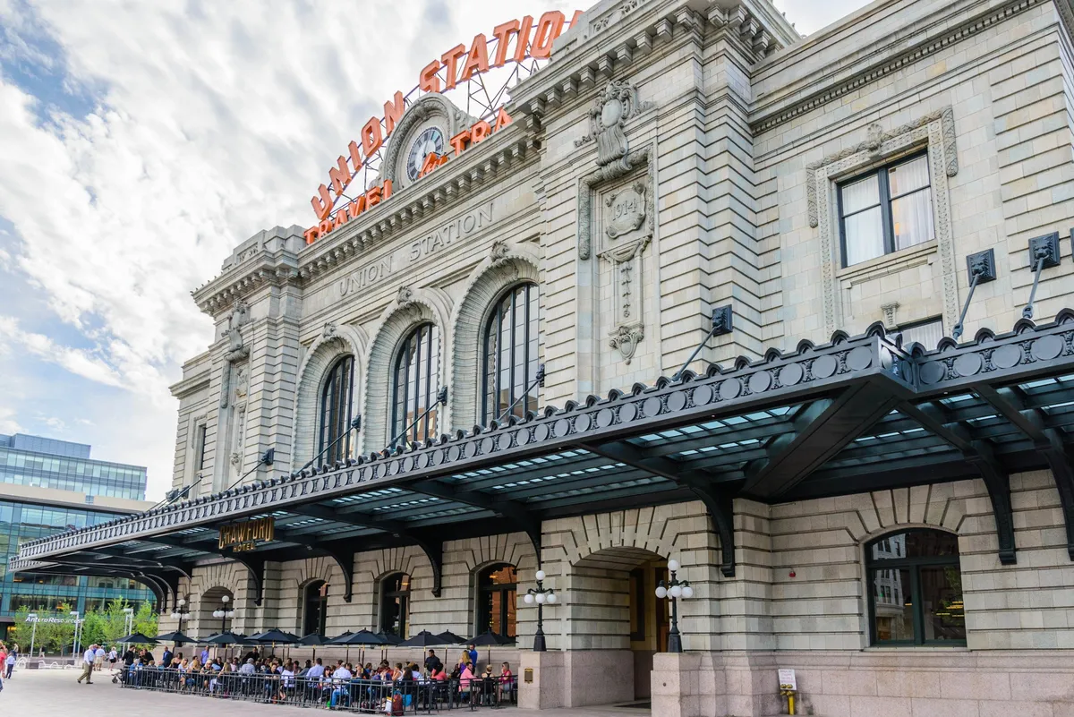 A bustling scene at Denver's iconic Union Station, featuring the recognizable 'Union Station' sign, architectural details, and people enjoying meals outside. The area is lively with social gatherings and travelers, emphasizing the station's role as a major transportation hub.