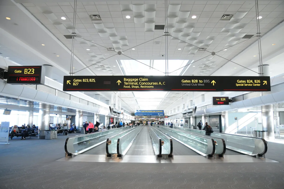 The image depicts a dynamic airport terminal with moving walkways, prominent gate signs, seating areas, and people engaged in activities such as traveling, shopping, waiting for flights, and eating at restaurants. The environment is bustling and lively, capturing the sense of movement and anticipation often associated with travel and new destinations.
