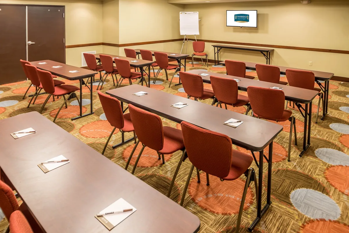 A bright, collaborative workspace with rows of tables and red chairs, set up for meetings, workshops, or focused work sessions. A presentation screen and whiteboard suggest a productive environment ideal for business travelers or guests needing a laptop-friendly area.